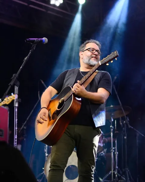 Cantor de óculos e barba tocando violão no palco, sob feixes de luz azul